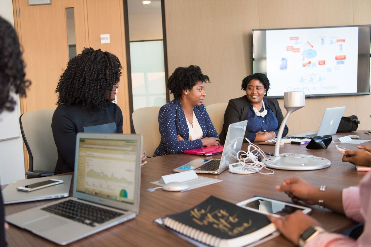 Colleagues gathered around a conference table with laptops, discussing a presentation on a wall screen