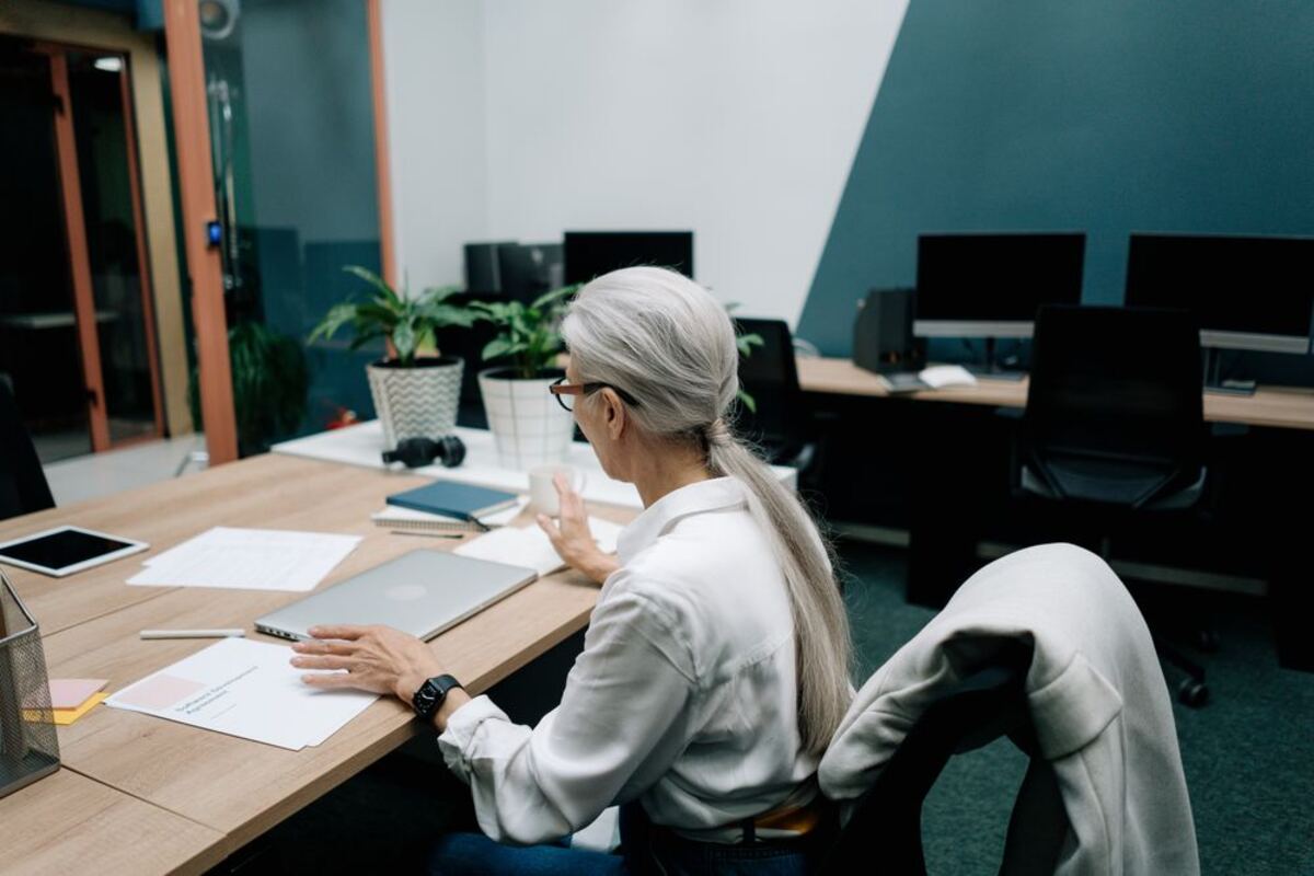 A woman with long gray hair works independently at a desk with her laptop and notes