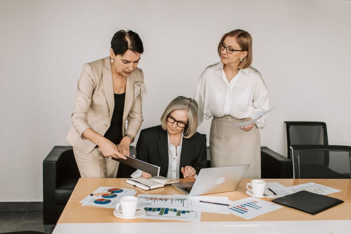 Three women in business attire review charts and a laptop together at a conference table